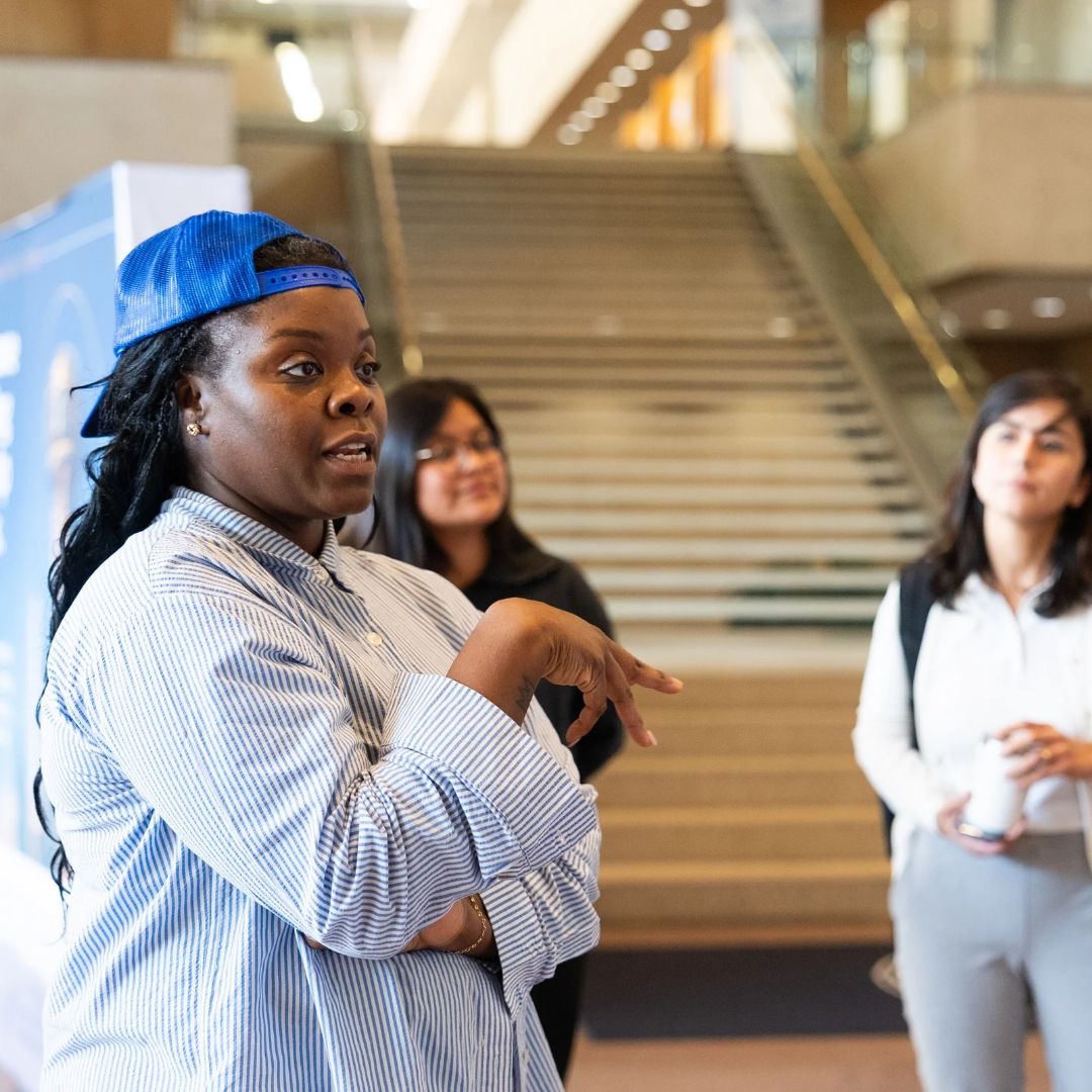 Feleceia Wilson speaking with participants in a public indoor space, wearing a blue cap and striped shirt.